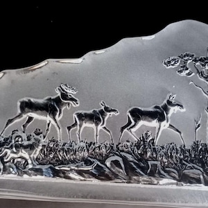 May include: A clear glass paperweight featuring an etched design of four deer in a woodland scene. The deer are arranged in a line, with the largest deer at the front and the smallest at the back. The woodland scene includes trees and shrubs.
