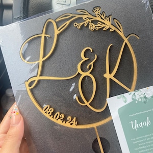 Könnte beinhalten: Ein goldener Holz-Kuchentopper mit den Buchstaben L & K in einer kursiven Schrift, umgeben von einem floralen Kranz. Das Datum 08.02.24 ist unter den Buchstaben eingraviert.