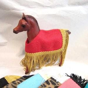 May include: A red felt horse blanket with gold fringe trim. The blanket is draped over a brown toy horse.