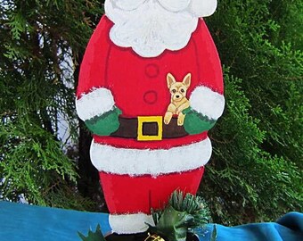 May include: A wooden Santa Claus figure with a small dog in his arms, painted in red, white, and green. The figure has a white beard and a red hat. The base features a pine cone, green apple, and a small green drum. The figure is approximately 12 inches tall.