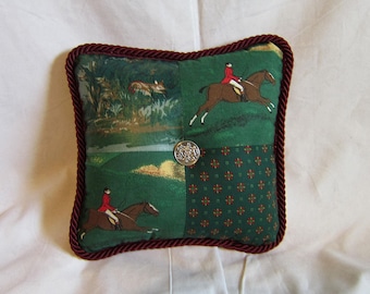 May include: A green and red patterned throw pillow with a burgundy rope trim and a silver button in the center. The pillow features a repeating pattern of small red and green flowers and a scene of a rider on horseback.