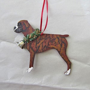 May include: Wooden Boxer dog ornament with a red ribbon for hanging. The dog is painted with brown, black, and white details, and wears a green garland with a silver bell. A festive decoration for dog lovers.