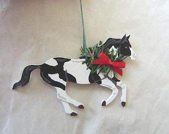 Könnte beinhalten: Ein schwarz-weiß bemaltes Holzpferde-Ornament mit einem grünen und roten Kranz um den Hals. Das Pferd rennt und hat eine grüne Schnur an seinem Kopf zum Aufhängen.