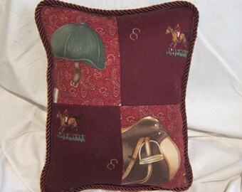 May include: A burgundy pillow with a brown, white, and green equestrian themed design. The pillow has a brown cord trim and features a riding helmet, a saddle, and a horse rider.