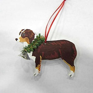 May include: Wooden dog ornament with a red ribbon for hanging. The dog is painted brown, white, and tan, with a green wreath around its neck and a silver bell. A festive decoration for the holidays.