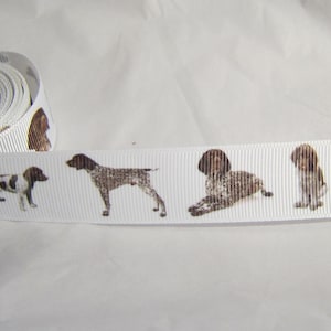 May include: White ribbon featuring a repeating pattern of brown and white dog illustrations. The ribbon is rolled up on the left side, with the patterned section visible. The dogs are depicted in various poses.