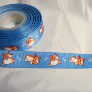 May include: A roll of blue ribbon featuring a repeating pattern of cartoon corgi dogs and white bones. The ribbon is made of a smooth, satin-like material, perfect for crafting and gift wrapping.