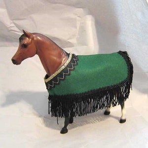 May include: A brown horse figurine wearing a green felt blanket with black fringe and a gold and black trim.