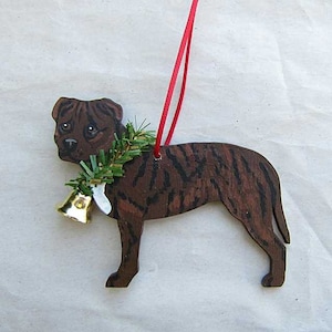 May include: Wooden dog ornament with a brown and black striped pattern. The dog has a green garland around its neck with a gold bell and a red ribbon for hanging. A festive holiday decoration.
