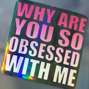 Könnte beinhalten: Ein rechteckiger Aufkleber mit dem Text "WHY ARE YOU SO OBSESSED WITH ME" in Rosa und Weiß. Der Aufkleber hat einen holografischen, irisierenden Hintergrund, der einen Regenbogen an Farben reflektiert.