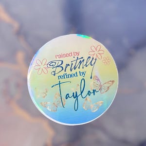 Puede incluir: Una pegatina redonda y holográfica con el texto "raised by Britney refined by Taylor". Presenta mariposas doradas y motivos florales rosas. El fondo tiene un efecto suave y borroso con tonos azules y dorados.
