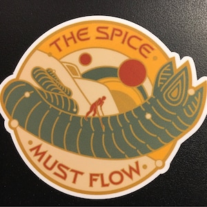 May include: A yellow and orange circular sticker with the text "The Spice Must Flow" in a white banner. The sticker features a green worm-like creature with a person in the background.