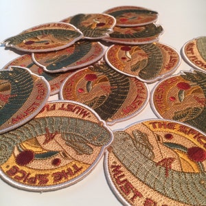 May include: A pile of embroidered patches with a variety of colours and designs. The patches feature a stylised image of a hand holding a red apple with the text "The Spice Must Flow" on the bottom.