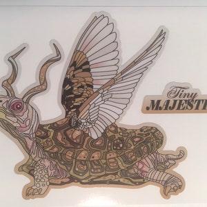 May include: A whimsical illustration of a turtle with wings and antlers. The turtle is brown and green with a detailed shell. The wings are white and gray with brown markings. The text "Tiny MAJESTIC" is in the upper right corner of the image.