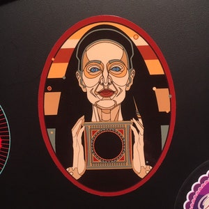 May include: A colorful illustration of a woman wearing a black and orange robe, holding a box with a black circle in the center. The woman has blue eyes and a red mouth. The background is a black oval with a red border.