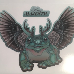 May include: A teal green frog with antlers and wings. The frog has a toothy grin and is sitting on a white background. The text "Tiny Majestic" is written above the frog.