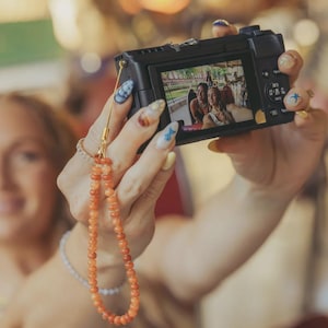 May include: A black digital camera featuring a screen displaying a photograph of two individuals. Attached to the camera is a wrist strap composed of small, round, orange-toned beads connected by a thin gold-colored cord and a metal loop.