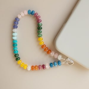 May include: A beaded phone strap with a rainbow of colors, including pink, blue, green, yellow, and orange. The strap is attached to a white phone case with a silver-colored clasp.
