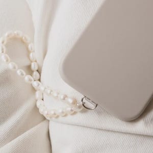 May include: A light gray phone case with a pearl wrist strap rests on white denim. The phone case has a camera module with three lenses. The pearl strap is made of small, round, white pearls and a silver-colored clasp.