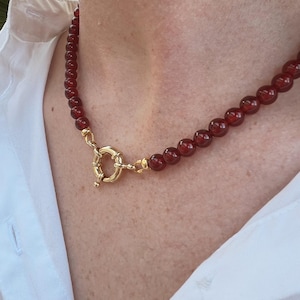 May include: A red gemstone necklace with a gold clasp. The necklace is made of small, round beads and has a simple, elegant design.