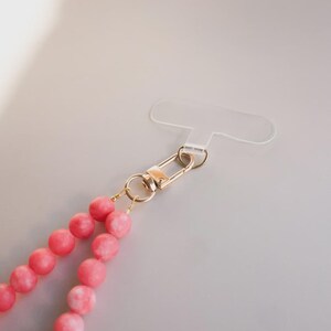 May include: A pink beaded phone strap with a gold-colored clasp and a clear plastic phone holder. The beads are a light pink color with white marbling. The clasp is a safety pin style.