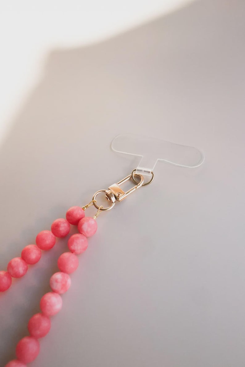 May include: A pink beaded phone strap with a gold-colored clasp and a clear plastic phone holder. The beads are a light pink color with white marbling. The clasp is a safety pin style.