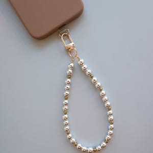 May include: A phone case in a brown hue is attached to a gold-colored clasp, which connects to a wrist strap. The strap is made of alternating silver and gold-colored beads, creating a stylish accessory.
