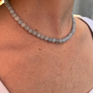Op de afbeelding: Een lichtblauwe kralenketting gemaakt van ronde, gladde stenen. De ketting wordt door een persoon gedragen, maar de focus ligt op de ketting zelf.