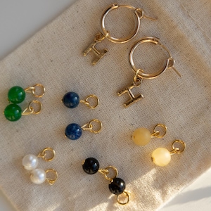 Puede incluir: Pendientes dorados con detalles de piedras preciosas y perlas. Incluyen aros con colgantes de letras y pendientes colgantes más pequeños con cuentas verdes, azules, negras y amarillas. Se exhiben sobre una tela beige.