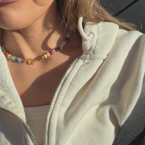 May include: A colorful beaded necklace with a gold clasp. The necklace features a variety of beads in shades of red, yellow, green, blue, and white. The necklace is worn with a cream-colored zip-up hoodie.