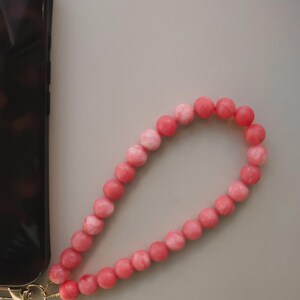 May include: A beaded phone wrist strap in shades of pink. The strap is made of round, polished beads and attached to a gold-colored clasp. The strap is next to a phone with a tortoiseshell case.