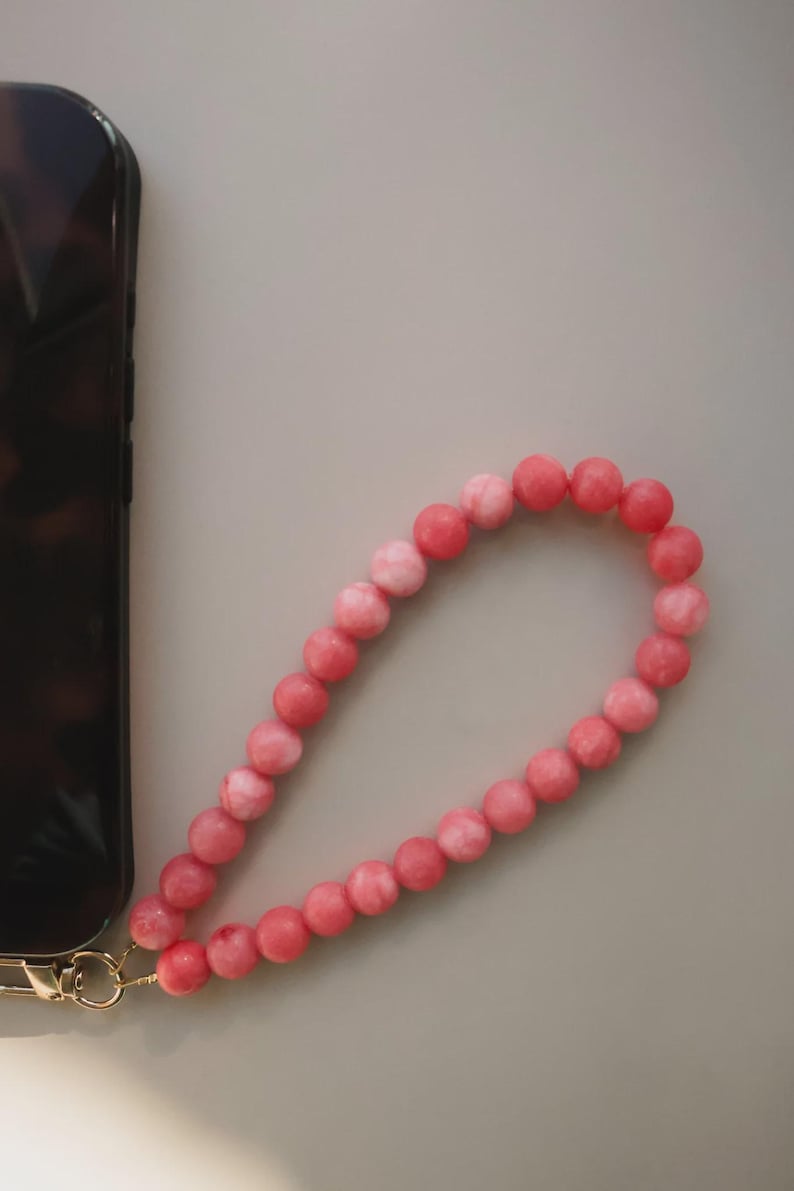 May include: A beaded phone wrist strap in shades of pink. The strap is made of round, polished beads and attached to a gold-colored clasp. The strap is next to a phone with a tortoiseshell case.