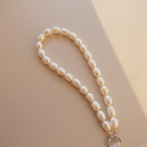 K&ouml;nnte beinhalten: Ein wei&szlig;es Perlenarmband mit einem silbernen Verschluss. Die oval geformten Perlen sind aufgereiht. Das Armband wird auf einer hellbeige Oberfl&auml;che pr&auml;sentiert.