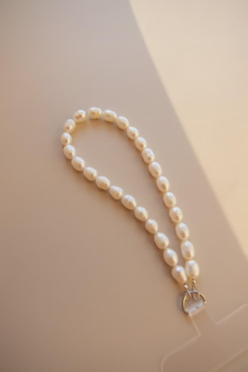 K&ouml;nnte beinhalten: Ein wei&szlig;es Perlenarmband mit einem silbernen Verschluss. Die oval geformten Perlen sind aufgereiht. Das Armband wird auf einer hellbeige Oberfl&auml;che pr&auml;sentiert.