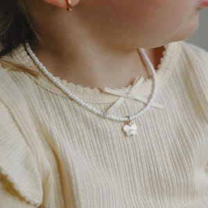 May include: A delicate pearl necklace with a butterfly pendant. The necklace is worn over a light yellow ribbed shirt. The butterfly pendant is white and hangs from a gold-colored ring. The shirt has a small bow detail.