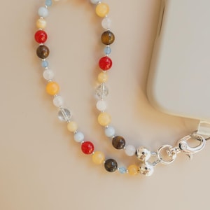 May include: A beaded phone strap featuring a variety of colorful gemstone beads, including red, yellow, brown, and clear quartz. The strap is attached to a silver-colored clasp and is designed to be attached to a phone case.