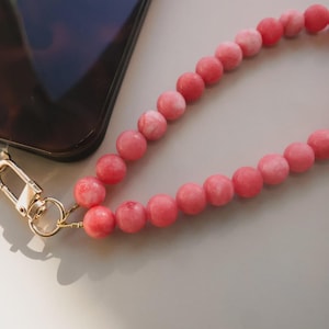 May include: A close-up of a phone with a tortoiseshell pattern case and a pink beaded wrist strap. The strap is made of round, pink beads and has a gold-colored clasp.