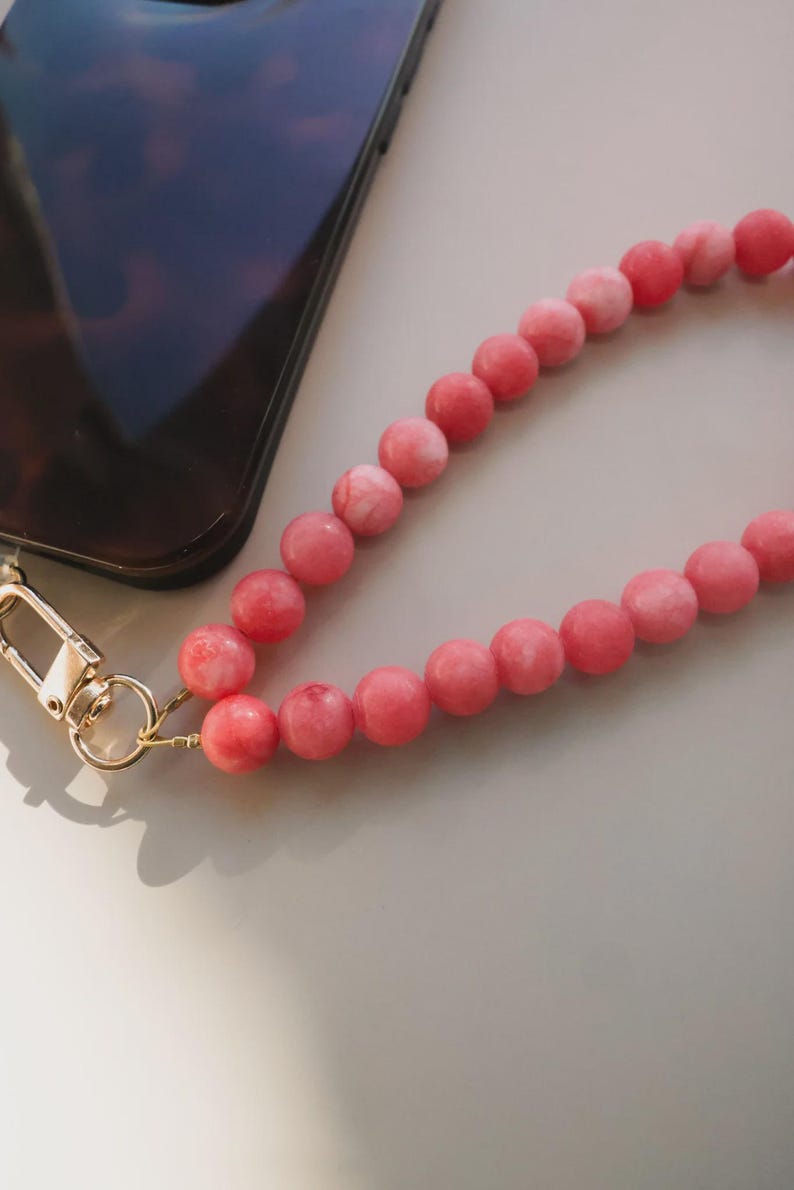 May include: A close-up of a phone with a tortoiseshell pattern case and a pink beaded wrist strap. The strap is made of round, pink beads and has a gold-colored clasp.