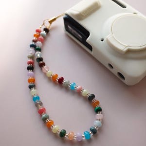 Peut inclure: Une dragonne de caméra perlée avec une variété de perles colorées dans les tons de rose, orange, vert, bleu et transparent. La dragonne est attachée à un étui d'appareil photo blanc avec un fermoir doré.