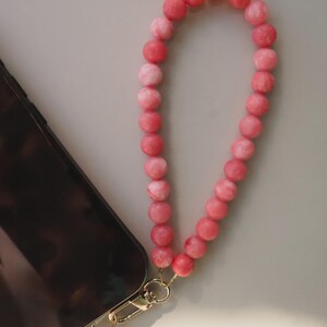 May include: A pink beaded phone wrist strap with a gold-colored clasp is attached to a black phone. The beads are a mix of light and dark pink. The phone is on a light gray surface.