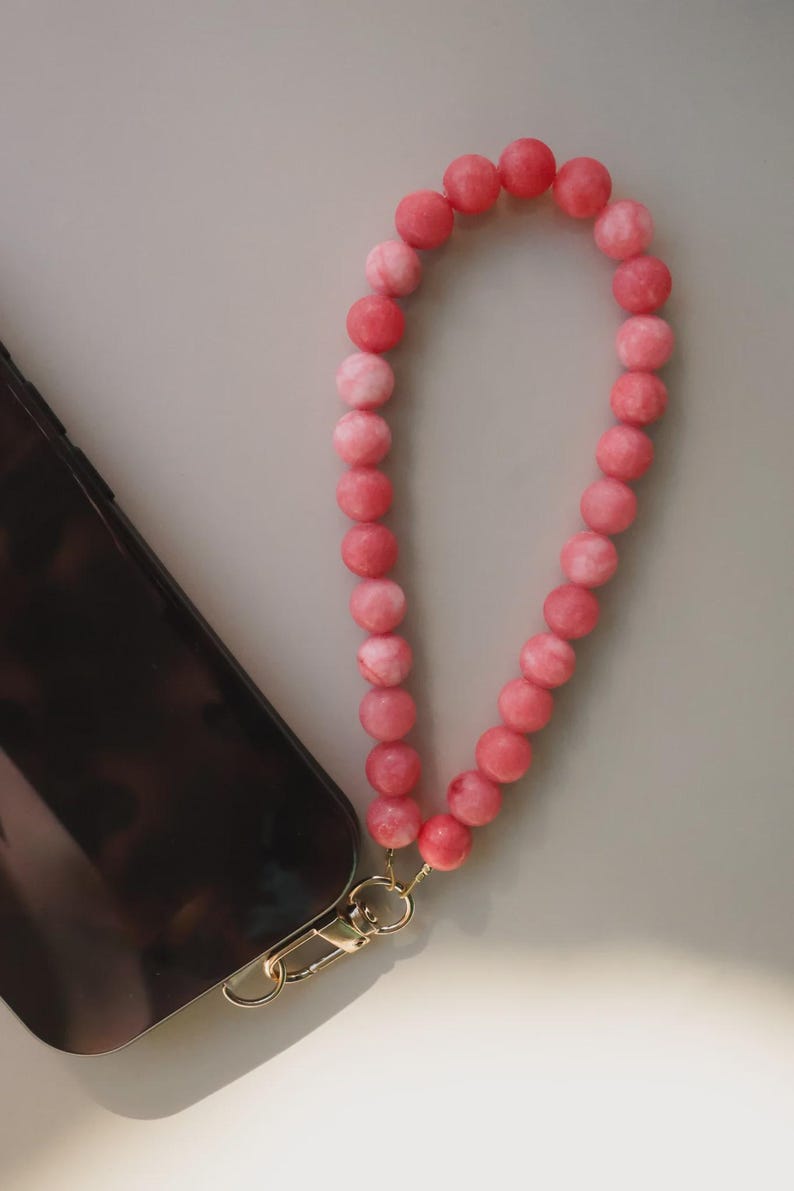 May include: A pink beaded phone wrist strap with a gold-colored clasp is attached to a black phone. The beads are a mix of light and dark pink. The phone is on a light gray surface.