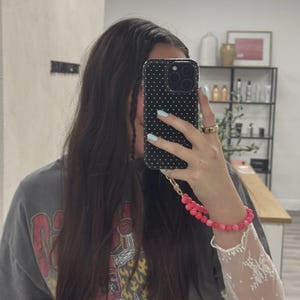 May include: A black phone case with white polka dots is held up in front of a person's face. A pink beaded bracelet and a white lace sleeve are visible. The person is wearing a gray t-shirt with a red graphic.