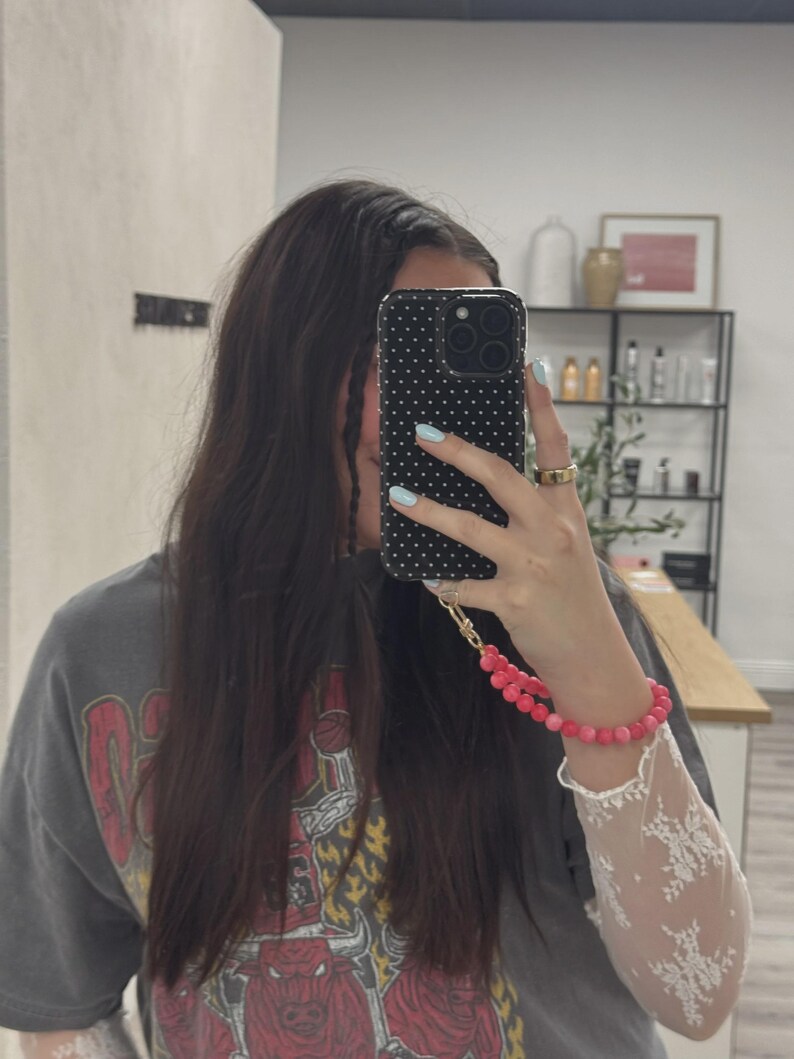 May include: A black phone case with white polka dots is held up in front of a person's face. A pink beaded bracelet and a white lace sleeve are visible. The person is wearing a gray t-shirt with a red graphic.