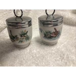 May include: Two white porcelain egg coddlers with silver-tone metal lids featuring ring handles. Each container displays a colorful illustration of a small bird perched among green pine needles or near water with reeds and aquatic plants.
