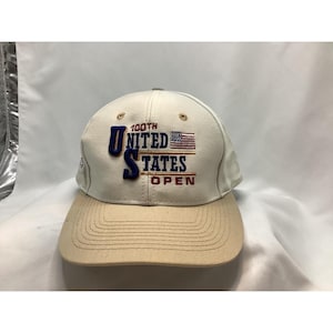Op de afbeelding: Crèmekleurige baseballcap met een bruin petje. De pet heeft geborduurde tekst en een vlagontwerp. De tekst luidt "100th UNITED STATES OPEN" in marineblauw en rood.