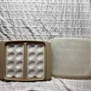 May include: A tan plastic egg container with two sections, each holding 12 egg-shaped indentations. The container has a matching lid. The egg holder is open, revealing the white interior. The lid is translucent.