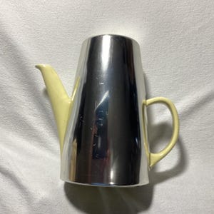 Könnte beinhalten: Eine hohe, zylindrische Kaffeekanne mit einem stark reflektierenden, polierten Chromkörper. Sie ist mit einem blassgelben Griff und einem passenden blassgelben Ausguss versehen, die beide eine glatte, glänzende Oberfläche aufweisen.