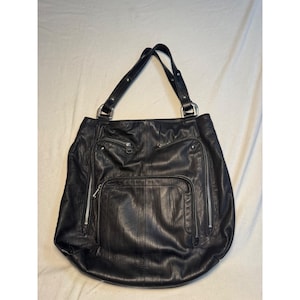 May include: A black leather tote bag featuring two shoulder straps with silver-tone metal hardware. The exterior includes two vertical zippered pockets on the sides and a large central zippered pocket, all finished with silver-tone metal zippers.