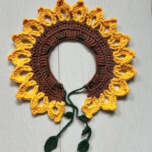 May include: A crocheted sunflower necklace with brown and yellow petals and green leaves. The necklace is tied with a brown cord.