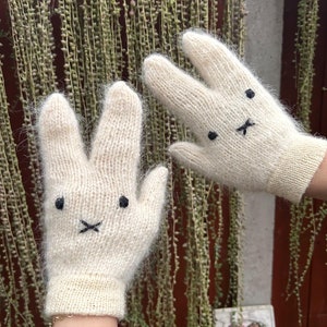 Op de afbeelding: Een paar witte gebreide wanten met konijnengezichten. De wanten hebben zwarte geborduurde ogen, een neus en een mond.